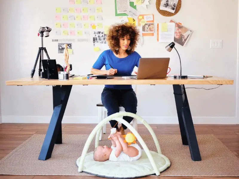 Junge Mutter arbeitet im Homeoffice am Laptop, während ihr Baby unter dem Schreibtisch spielt – Symbol für Vereinbarkeit von Beruf und Familie